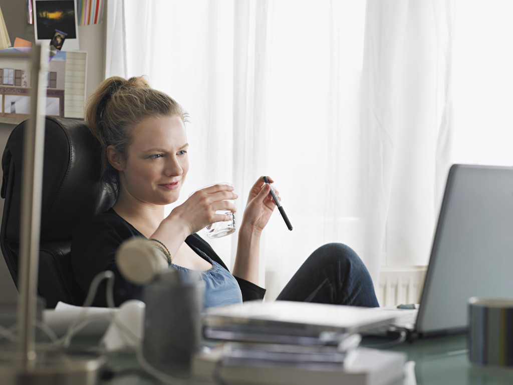 Young woman holding water glass while looking at laptop in home office
