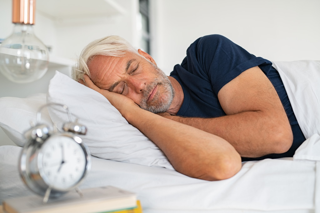Mature man sleeping on a bed at home with an alarm clock on a bedside table.